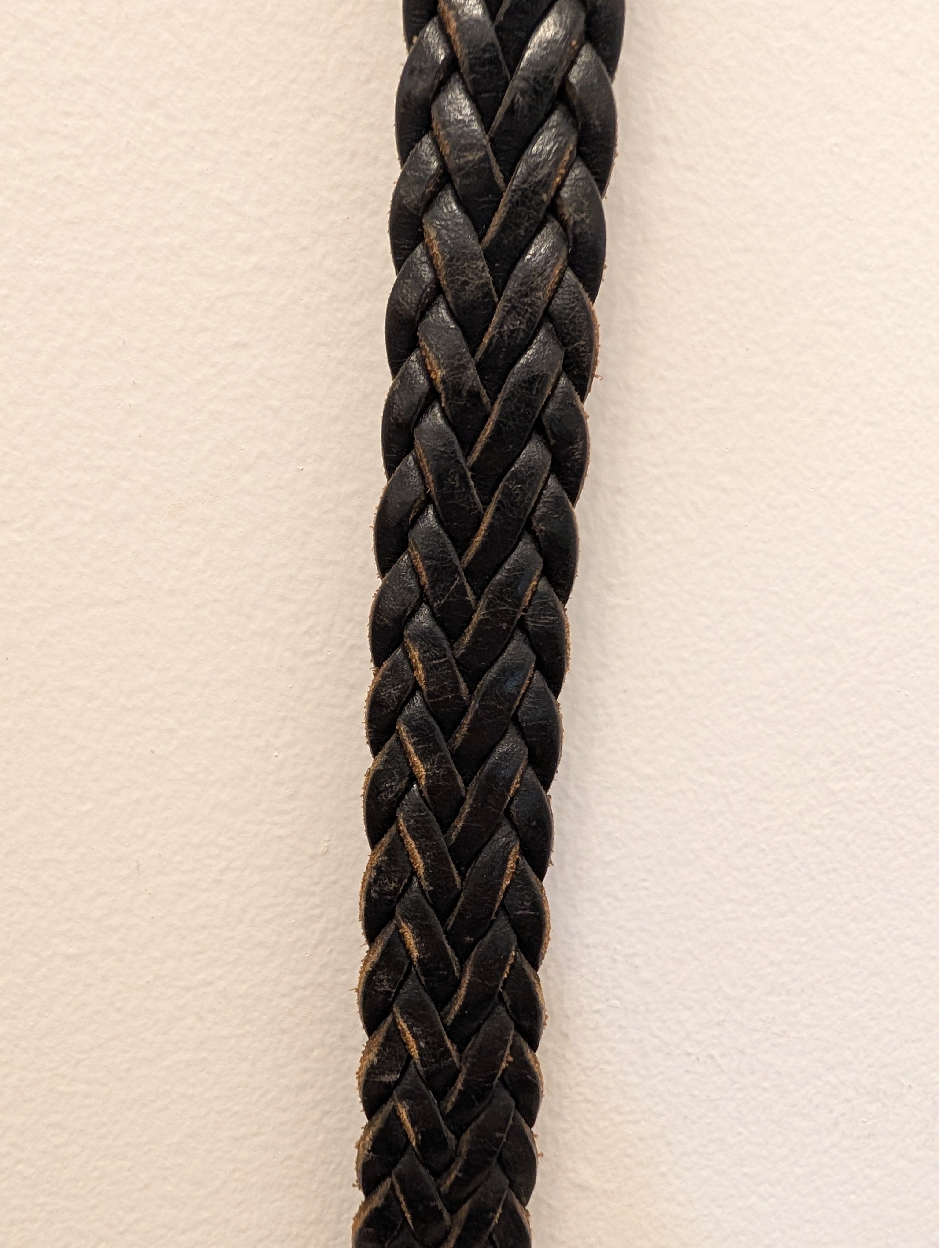 A close-up of a thick black braided leather strap or cord, vertically oriented against a plain off-white background.