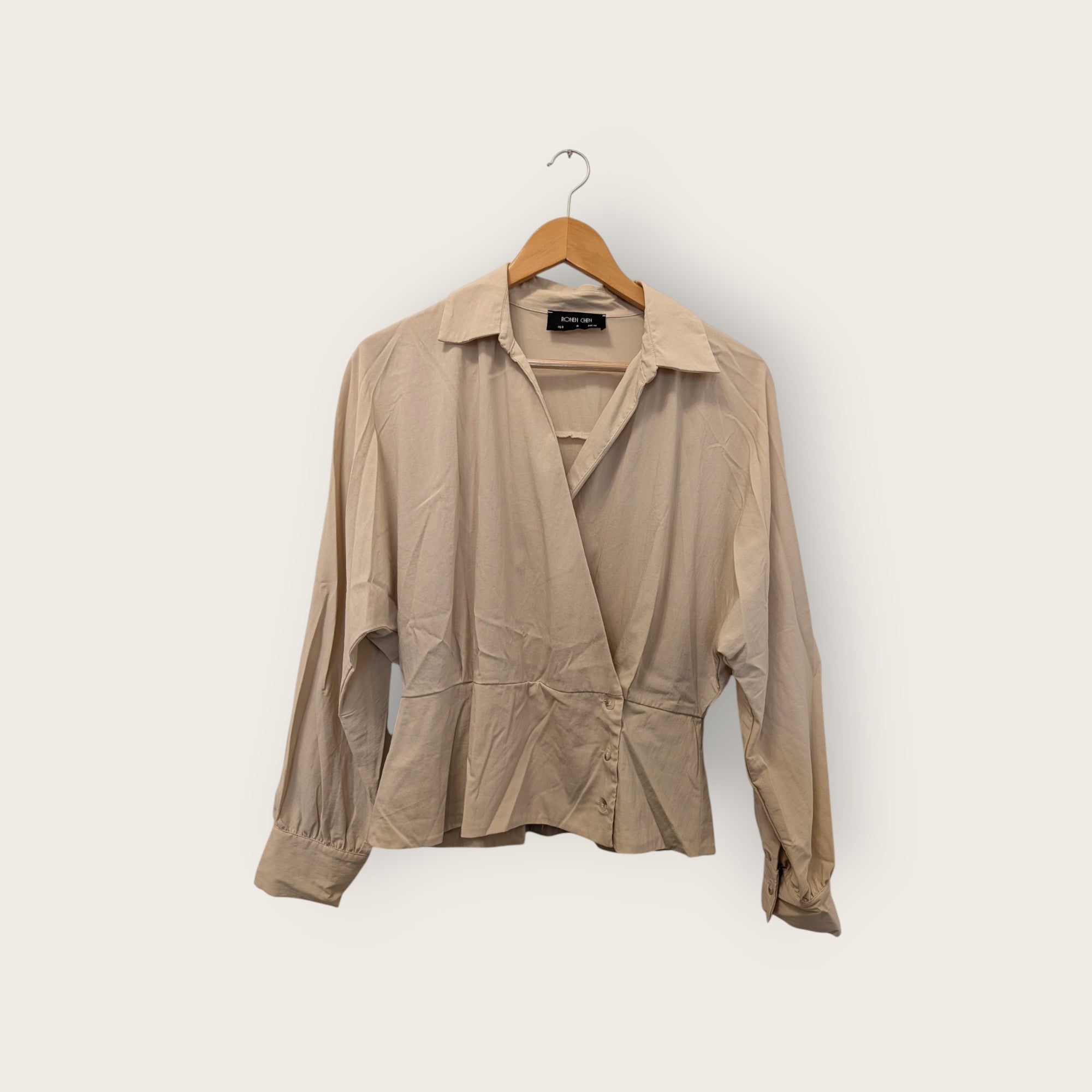 A beige long-sleeve wrap blouse with a pointed collar and front button closure, hanging on a wooden hanger against a plain off-white background.