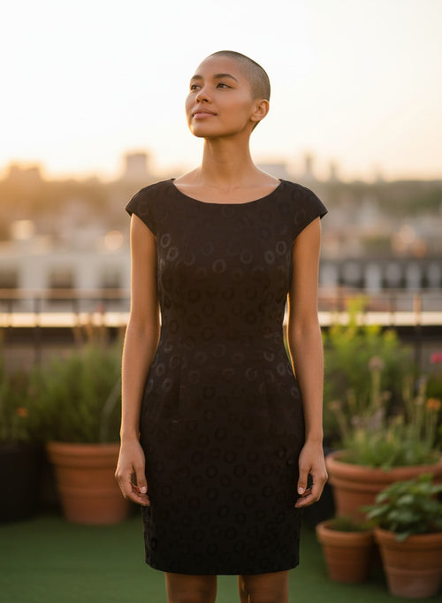 Model wearing a black Nu-Nv Clothing cap sleeve sheath dress with tonal circle jacquard pattern on a rooftop.