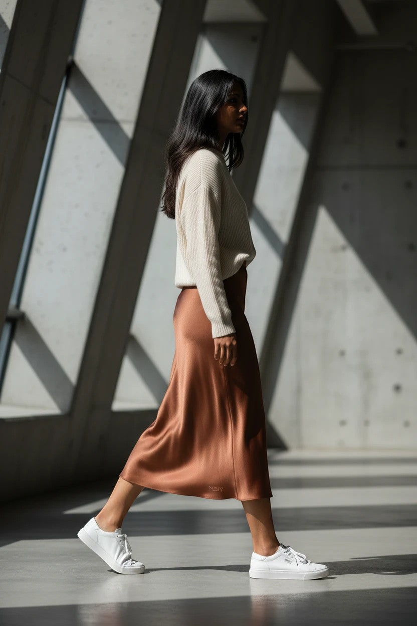 Model wearing a Nu-Nv Clothing brown satin midi skirt with a cream sweater and white sneakers in a modern space.