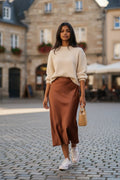 Model wearing a Nu-Nv Clothing brown satin midi skirt with a cream ribbed sweater and white sneakers in a city square.